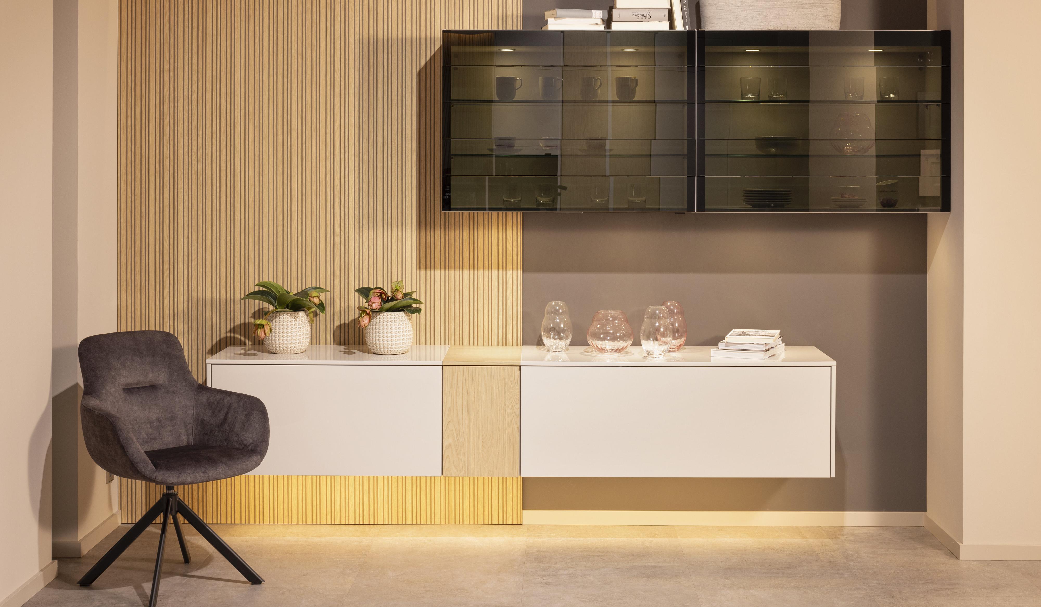 Wandmontiertes Lowboard in Weiss mit Holzeinsatz mittig schräg darüber schwarzer Hänge-Vitrinenschrank davor dunkelgrauer Samt-Polster-Drehstuhl